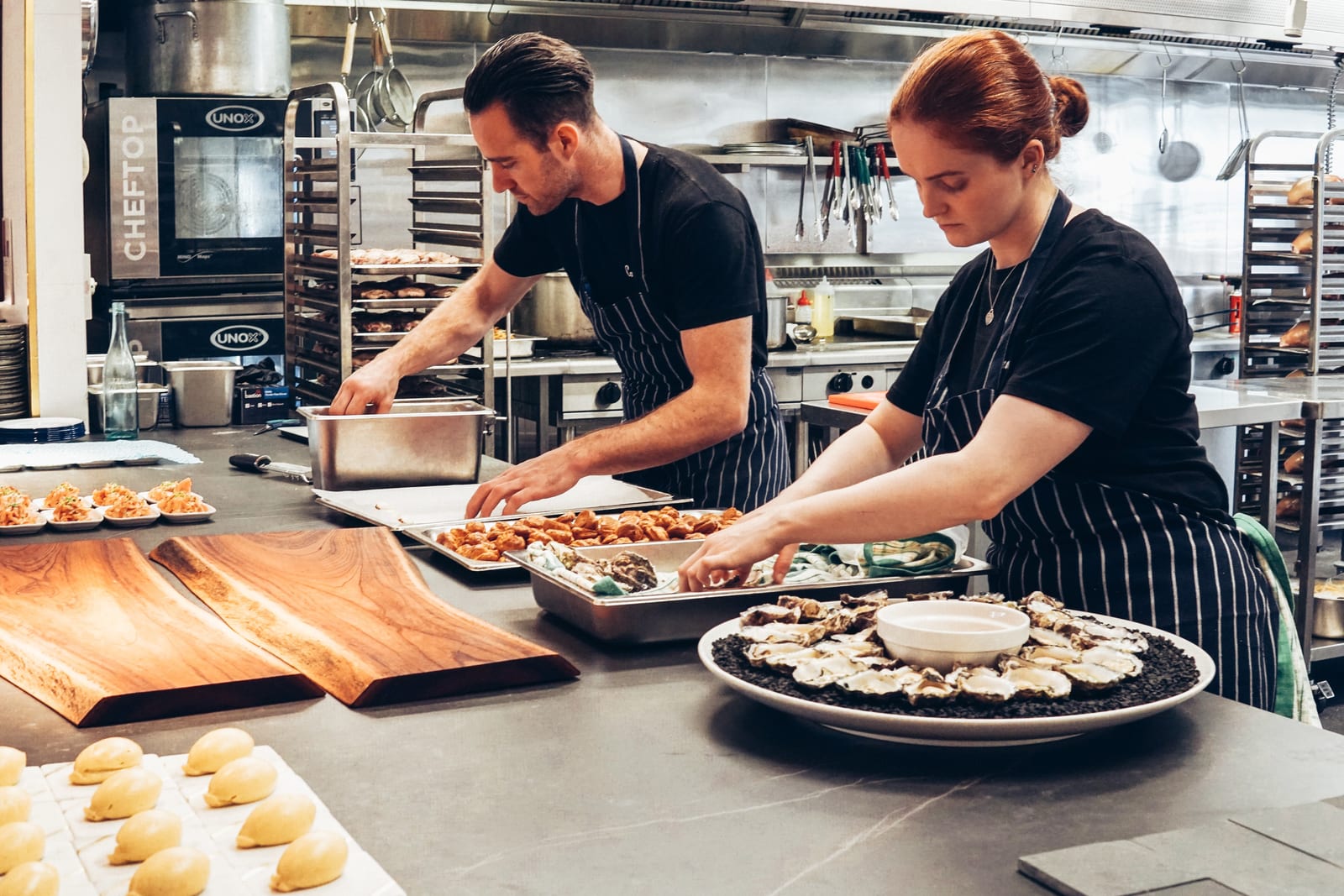 Deux cuisiniers dressant des plats, bras tendus au-dessus d'un plan de travail en inox